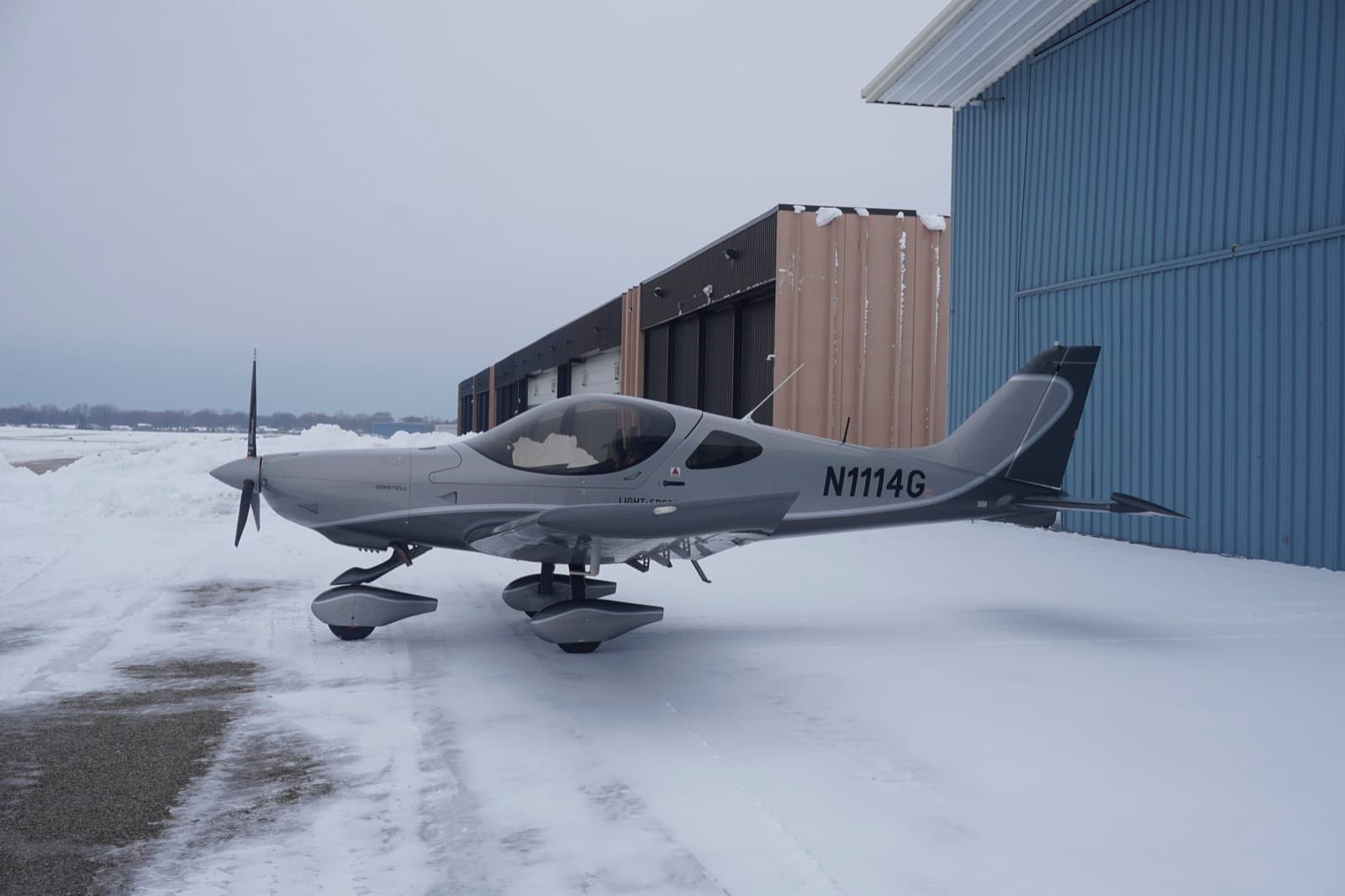 The silver Bristell N1114G parked on a snow covered ramp beside the hangar